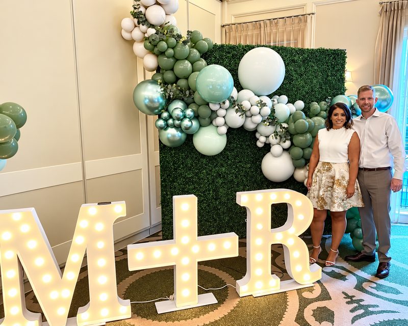 Couple beside a green and white balloon garland and lit marquee letters M + R